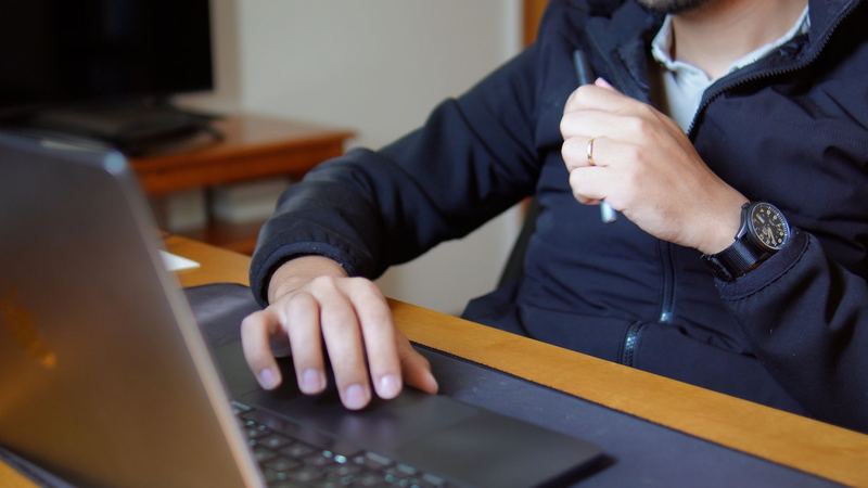João à mesa com caneta na mão e relógio no pulso, ao lado do notebook